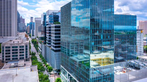 A photo of a large glassy skyrise in Midtown Atlanta under blue skies over a wide street with many buildings in the background and trees at the base.