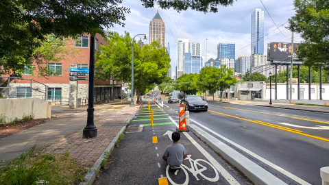 An image of a new two-way cycle track in Midtown Atlanta near many trees and tall buildings and cars, under gray-blue skies