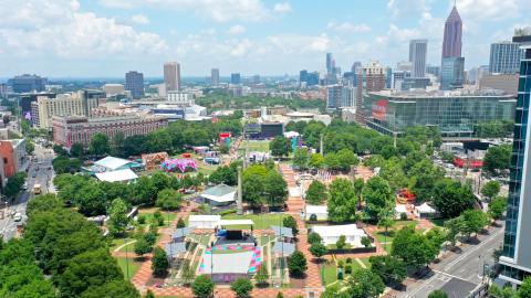 An aerial photo of a large city with a festival area in a park near many tall buildings under blue skies and many trees and wide street in downtown Atlanta.