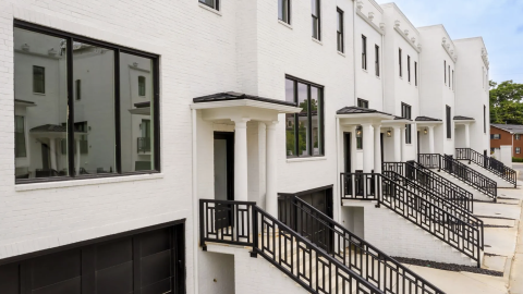 An image of a site with three-story white townhomes in brick with modern-style interiors near a wide street and many trees and large homes.